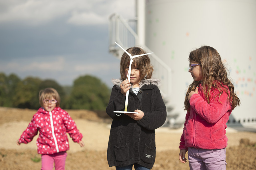 trois enfants portant une maquette d'éolienne avec éoliennes en fond